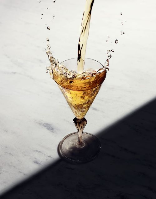 Pouring light-amber liquid into a stemmed martini glass on a marble surface, showcasing dynamic splashes and dark shadows, suggesting beverage advertisement.