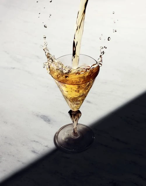 Pouring light-amber liquid into a stemmed martini glass on a marble surface, showcasing dynamic splashes and dark shadows, suggesting beverage advertisement.