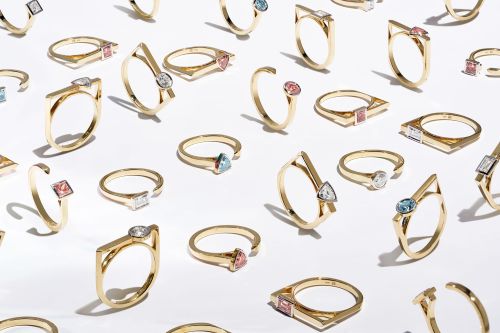 Collection of geometric rings in gold with colored stones arranged on a seamless white background, casting soft shadows.