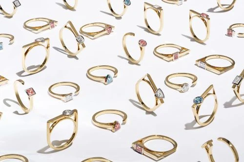Collection of geometric rings in gold with colored stones arranged on a seamless white background, casting soft shadows.