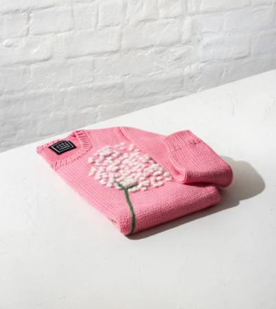 Folded pink knit sweater with a white floral appliqué and green stem resting on a white surface.