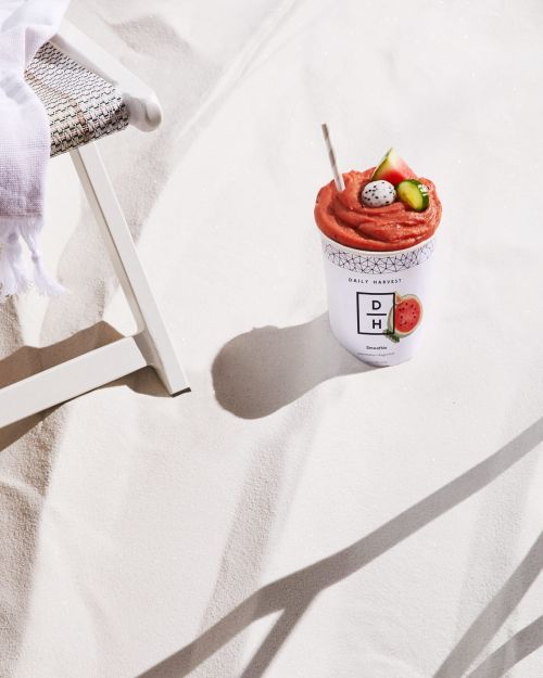 Fruit smoothie in a Daily Harvest container on a beach with a small, white chair in the background on a sunny day.