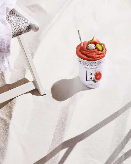 Fruit smoothie in a Daily Harvest container on a beach with a small, white chair in the background on a sunny day.