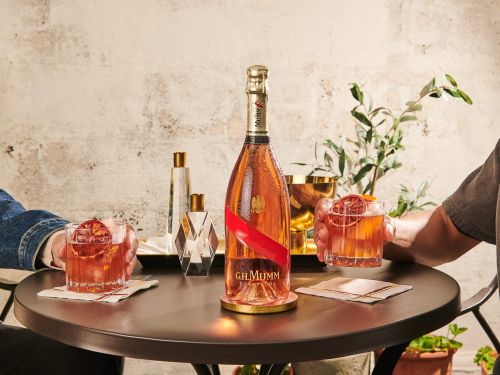 G.H. Mumm champagne bottle on a table next to cocktails with two people holding the drinks in a neutral-toned indoor setting.