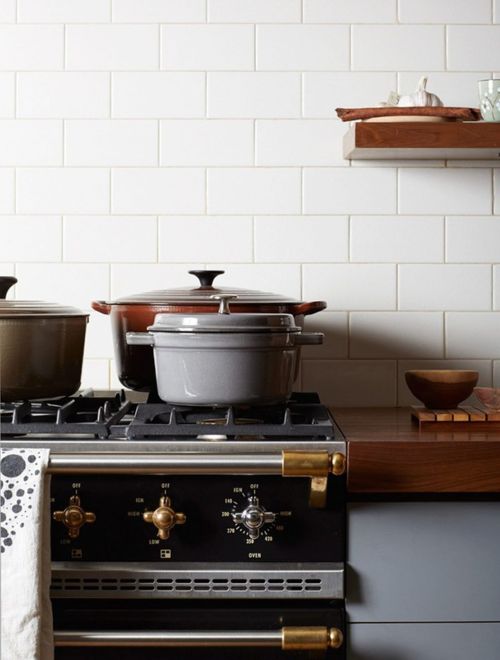 Still life of enameled cast iron cookware on a black gas range with brass fixtures in a bright kitchen with white subway tiles.