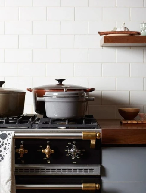 Still life of enameled cast iron cookware on a black gas range with brass fixtures in a bright kitchen with white subway tiles.