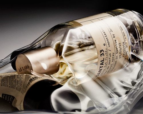 Close-up still life of a clear glass Le Labo perfume bottle with a metallic cap and paper label, wrapped in clear plastic.