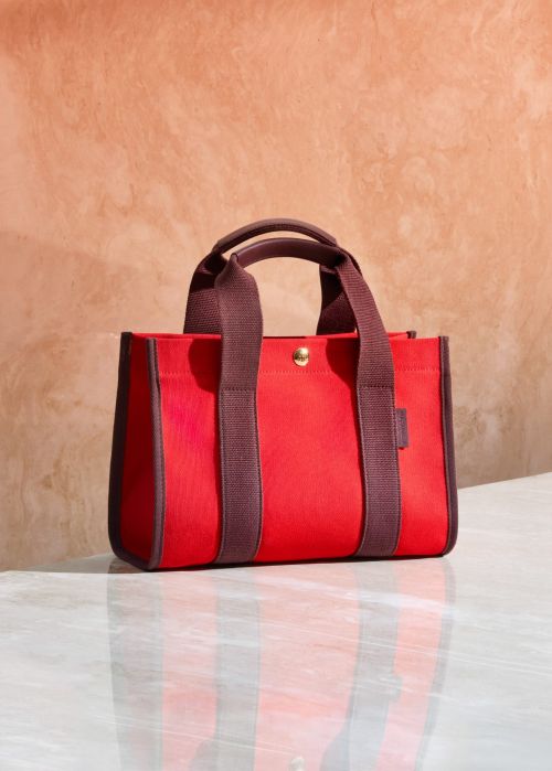 Red canvas tote bag with brown trim and handles on a marble surface in a brightly lit, neutral indoor setting.