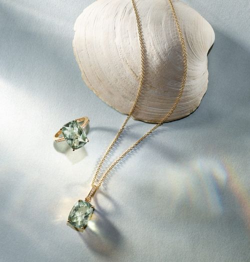 Elegant still life featuring a green amethyst pendant necklace, ring, and a textured seashell on a soft, light-blue surface.