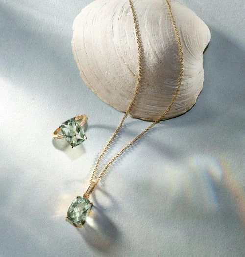 Elegant still life featuring a green amethyst pendant necklace, ring, and a textured seashell on a soft, light-blue surface.