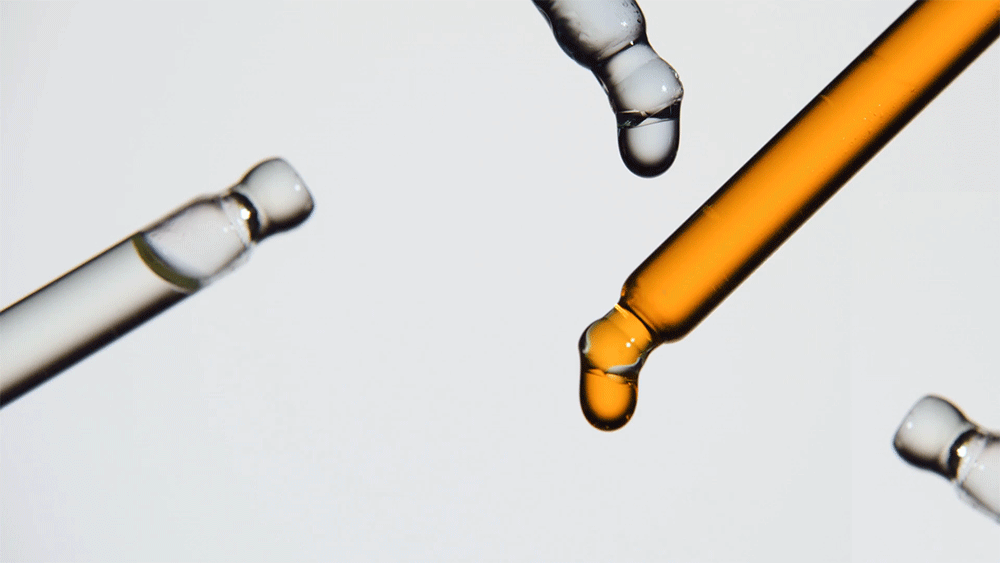 Macro photograph of multiple clear and amber glass droppers with hanging liquid droplets against a white background. Beauty product.