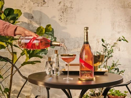 A person pours rose champagne into a gold-rimmed coupe glass on a round table with a bottle of champagne, a decanter, and plants.