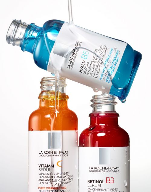 Three La Roche-Posay serum bottles, blue, orange, and red, in a studio shot with serum pouring from the blue bottle.