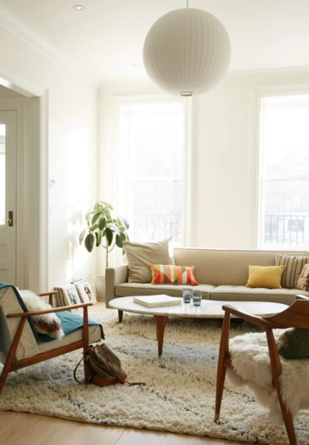 Living room interior featuring a beige sofa with pillows, a white round coffee table, wooden armchairs, a fluffy rug, and a globe pendant light.
