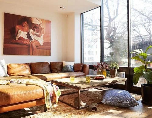 Sunny modern living room interior featuring a brown leather sectional sofa, a large painting, and floor-to-ceiling windows.