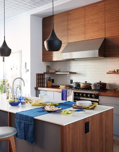 Modern kitchen interior showcasing an island, dark pendant lights, wood cabinets, stainless steel range hood, and various cookware.