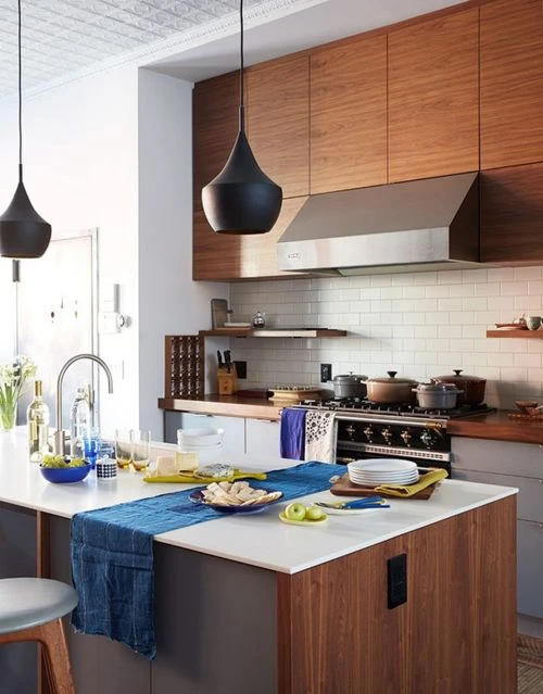 Modern kitchen interior showcasing an island, dark pendant lights, wood cabinets, stainless steel range hood, and various cookware.