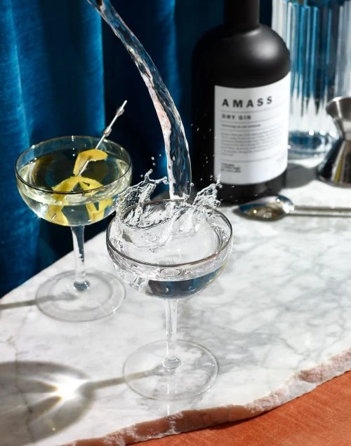 Crystal cocktail glasses filled with clear liquid and lemon twist, with Amass gin bottle, on marble surface, against blue curtain.