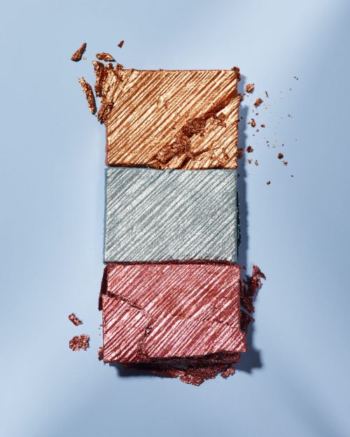 Three rectangular eyeshadow palettes stacked on top of each other in the colors of copper, silver, and rose-red makeup on a blue surface.