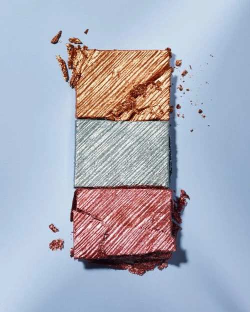 Three rectangular eyeshadow palettes stacked on top of each other in the colors of copper, silver, and rose-red makeup on a blue surface.