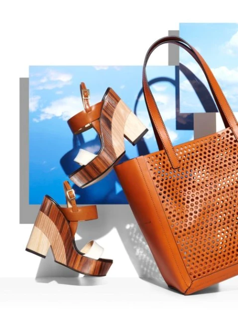 Studio shot of a wooden wedge sandals and a tan leather tote bag, positioned in front of a backdrop featuring blue sky and clouds.