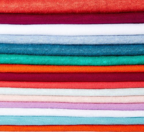 Stack of folded textile in orange, maroon, and blue tones forming a colorful, textured background. Fabric is closely arranged in layers.