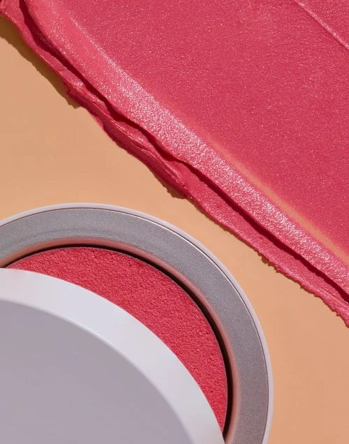 Overhead shot of a pink blush compact with the lid slightly open next to a smear of pink cream makeup on a peach-toned surface.