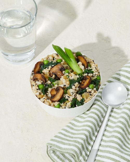 Overhead shot of a bowl filled with brown rice, mushrooms, peas, and green onions on a white textured surface next to a water glass.