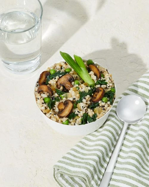 Overhead shot of a bowl filled with brown rice, mushrooms, peas, and green onions on a white textured surface next to a water glass.
