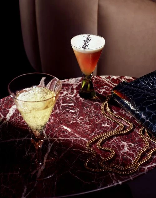 Two cocktails in martini glasses, one pink and the other yellow, sit on a marble table next to a gold chain and purse.