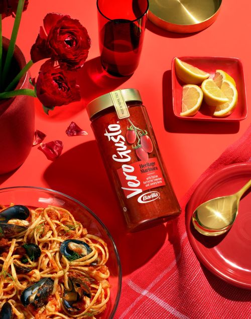 High-angle shot of marinara pasta with mussels, a jar of Vero Gusto sauce, lemons, flowers, and red accents creating a bold food still life.