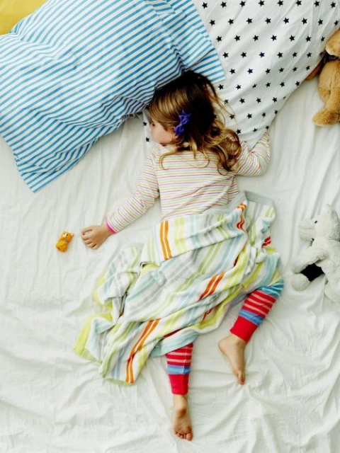 Top-down view of a young child asleep in bed with pillows and a colorful striped blanket, accompanied by stuffed animals and toys.