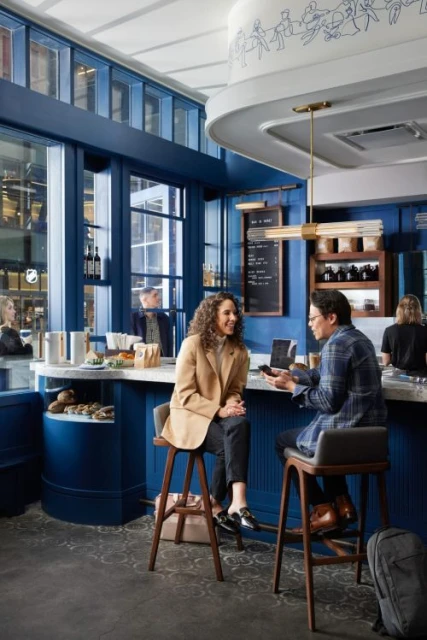 Woman in beige blazer seated across from a man at a cafe bar, with blue walls, wood stools, and modern lighting.