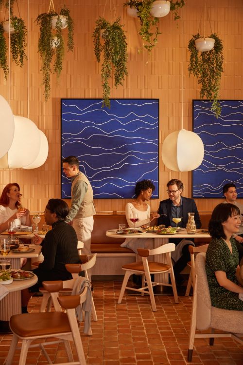 Indoor restaurant scene with several people seated at round tables enjoying food and drinks, under hanging plants and globe lights.