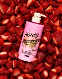 Pink body lotion bottle labelled Honey Strawberry resting among a dense bed of sliced and whole red strawberries.