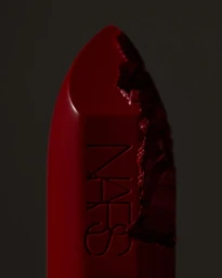 Macro shot of a vibrant red lipstick with an unevenly carved tip and embossed NARS logo against a dark background