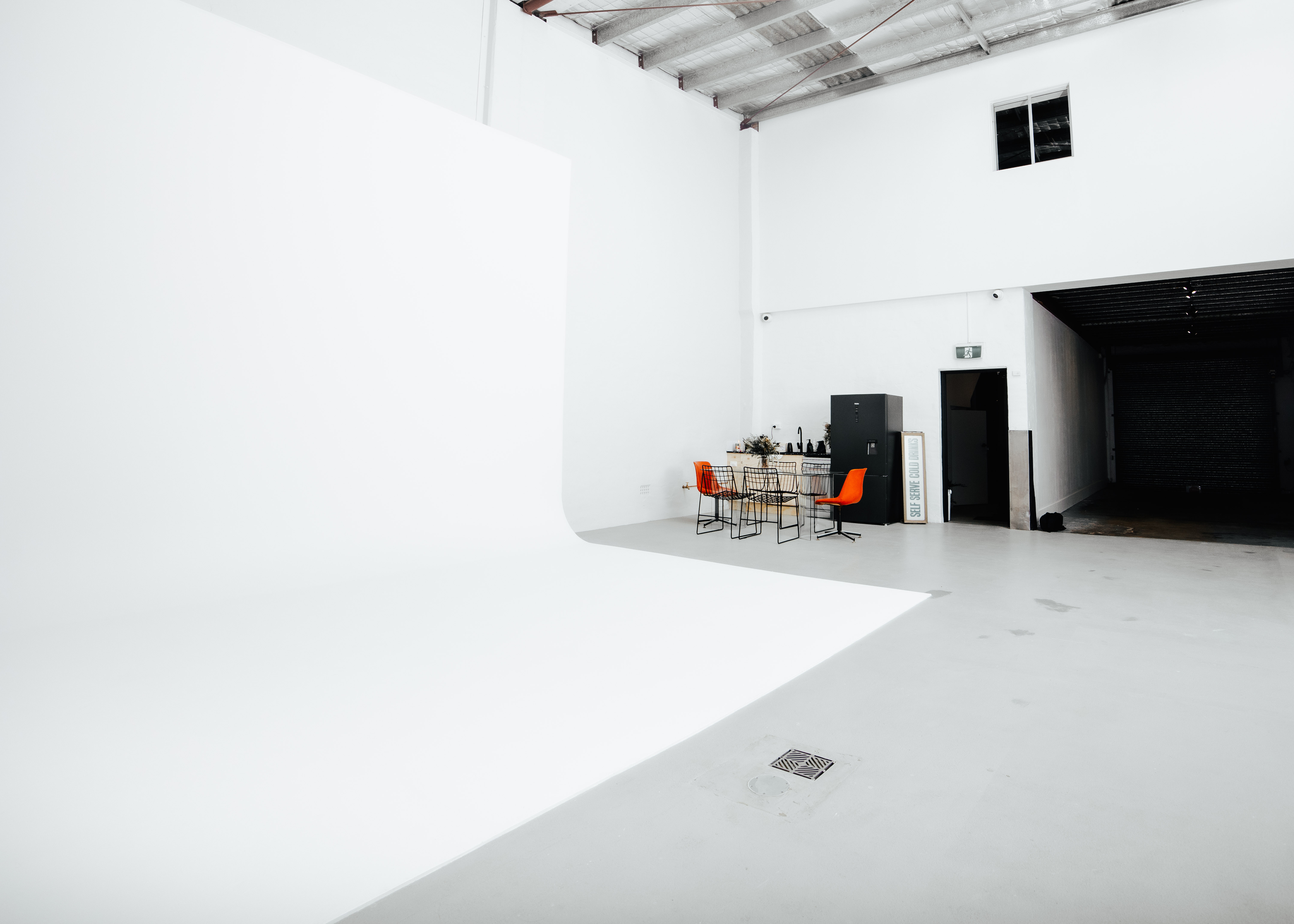 Interior view of a white photo studio with lighting equipment, orange chairs, a kitchen unit and a blank backdrop.