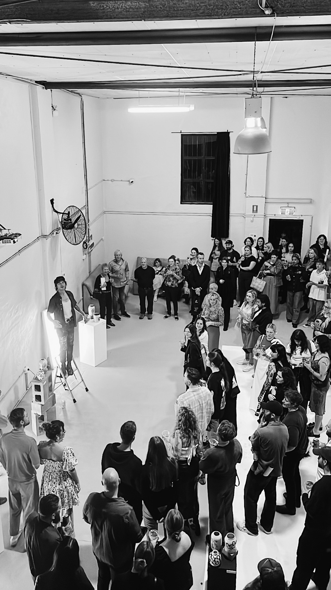 Monochrome shot of an event indoors featuring a group of people watching a speaker standing on cinder blocks, lit by spotlights.