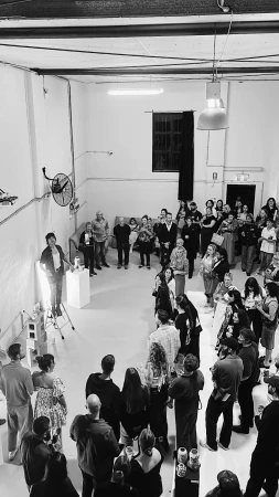 Monochrome shot of an event indoors featuring a group of people watching a speaker standing on cinder blocks, lit by spotlights.
