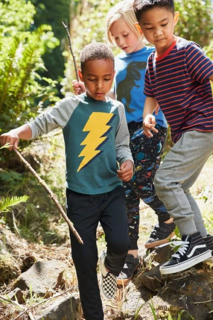 Three young boys hiking outdoors in a wooded area. The boy in front wears a green shirt with a yellow lightning bolt and black pants.