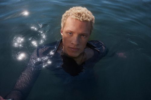 Adult male with blond hair stands waist-deep in dark water, wearing a black mesh long-sleeved shirt. The water reflects light.