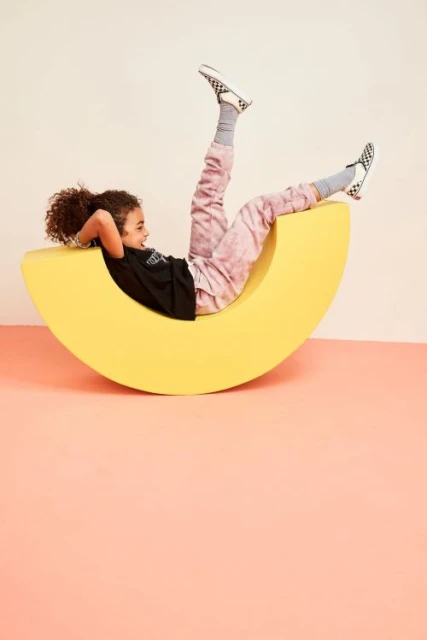 Child smiles while relaxing on a crescent moon shaped yellow prop, wearing a black t-shirt and tie-dye pants, indoor studio setting.