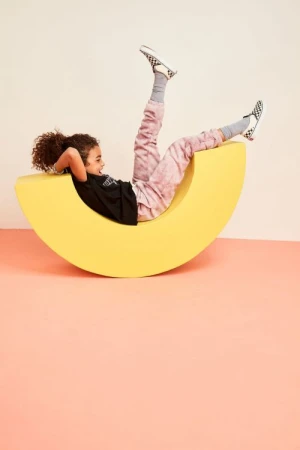 Child smiles while relaxing on a crescent moon shaped yellow prop, wearing a black t-shirt and tie-dye pants, indoor studio setting.