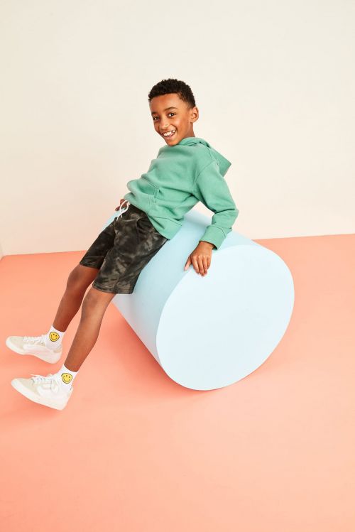 A young Black boy in a green hoodie and shorts smiles while leaning on a light blue cylinder, creating a happy and playful scene.