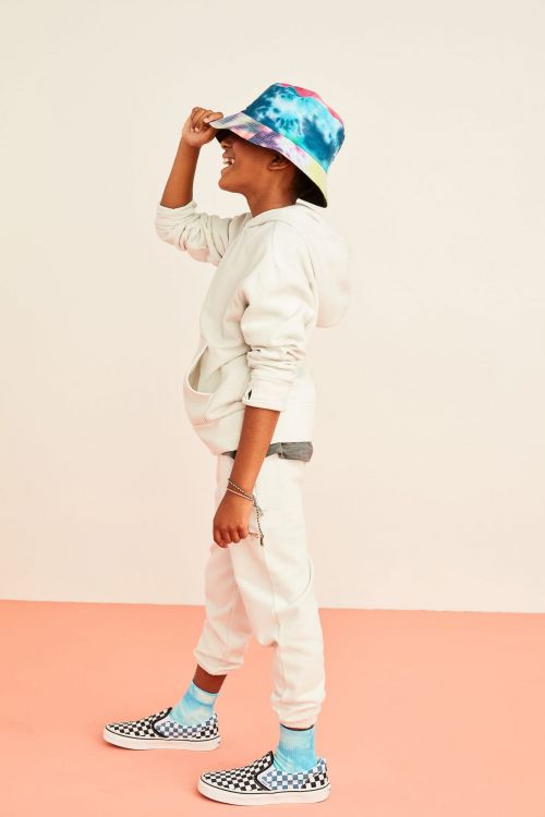 A child in a tie-dye bucket hat, hoodie, and sneakers smiles in a studio setting. The low-angle portrait captures youthful fashion.
