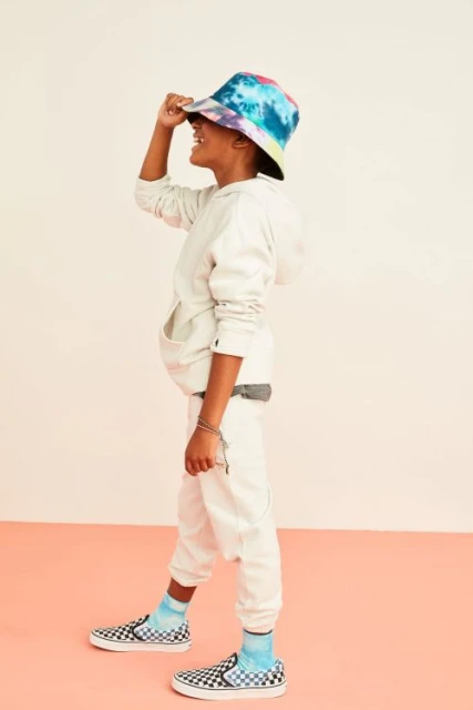 A child in a tie-dye bucket hat, hoodie, and sneakers smiles in a studio setting. The low-angle portrait captures youthful fashion.