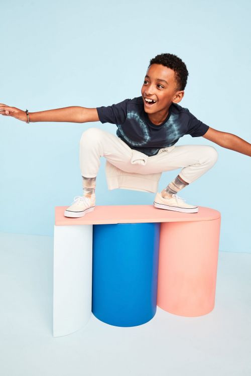 A boy is crouching on a three-column platform, posing in front of a light-blue backdrop in a studio environment with vibrant colors.