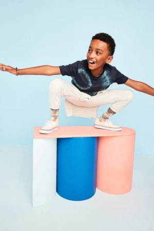 A boy is crouching on a three-column platform, posing in front of a light-blue backdrop in a studio environment with vibrant colors.