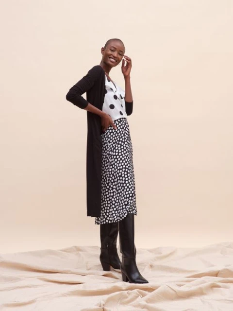 A Black woman stands smiling in a long black cardigan, black and white polka dot skirt and top, and black heeled boots on fabric.