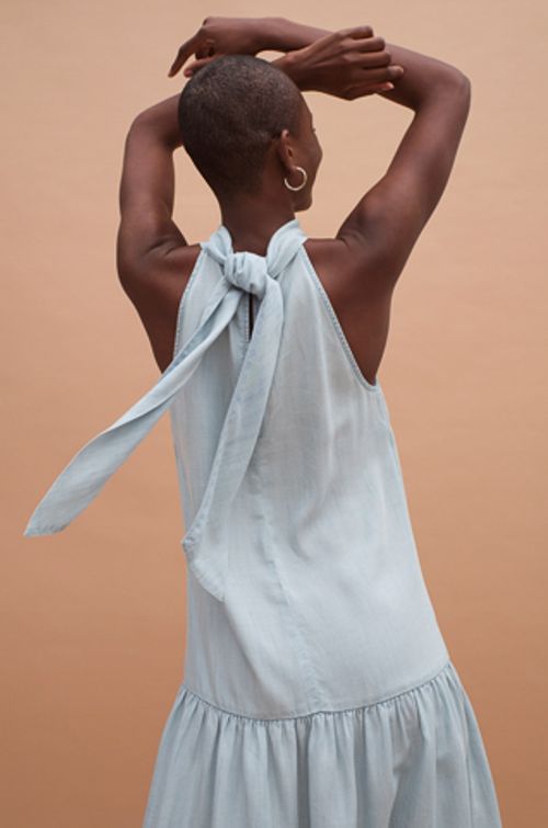 Back view of an adult woman with short hair wearing a light blue dress tied at the neck against a neutral brown background.